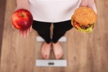 Voedingskeuzes: vrouw op een weegschaal met in de ene hand een appel en in de andere een hamburger.